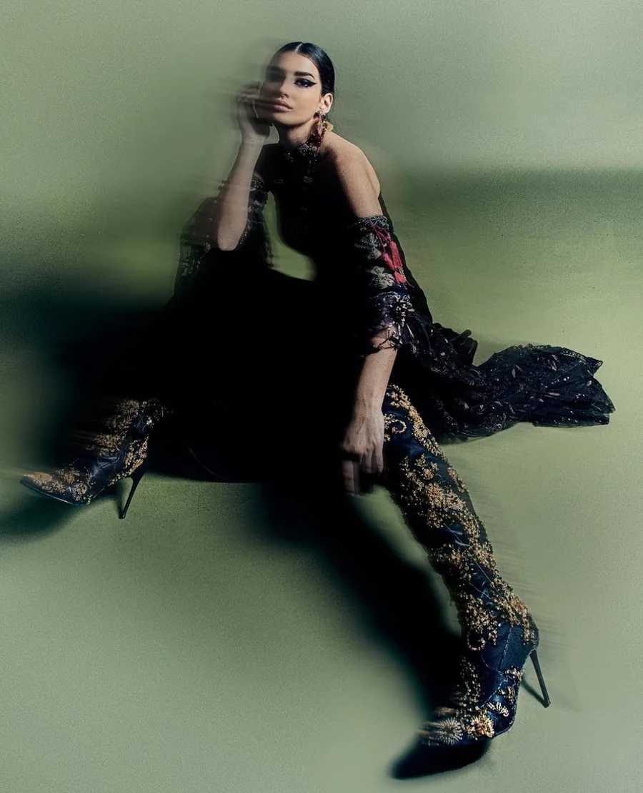 Model seated on a pale green floor in a dark lace dress and gold-embroidered thigh-high boots, hand at her cheek, with a motion-blurred edge.