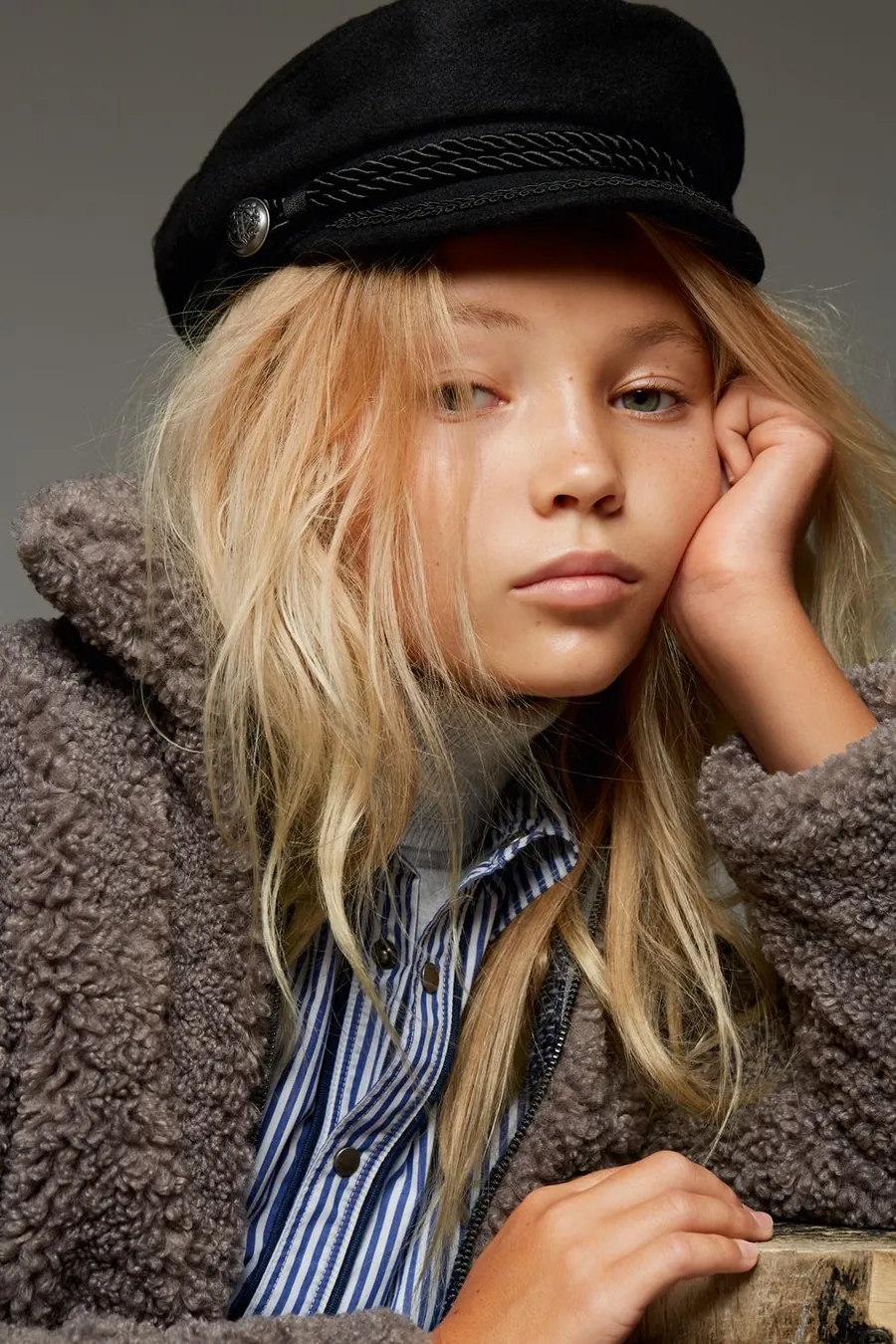 Girl with long blonde hair in a black cap and fur jacket, hand at her cheek, against a grey backdrop.
