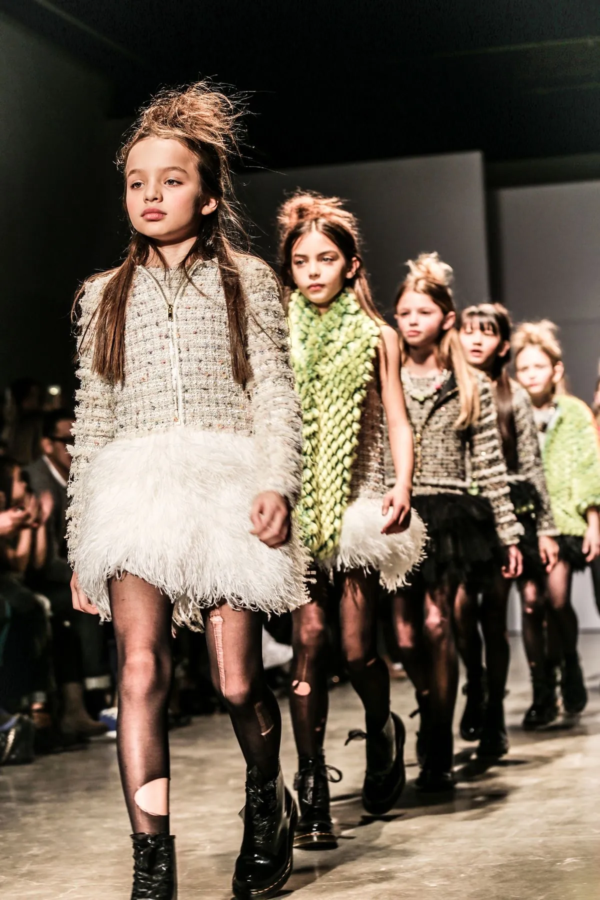 Girls walking the runway at a children's fashion show, in tweed jackets, feathered skirts and boots.