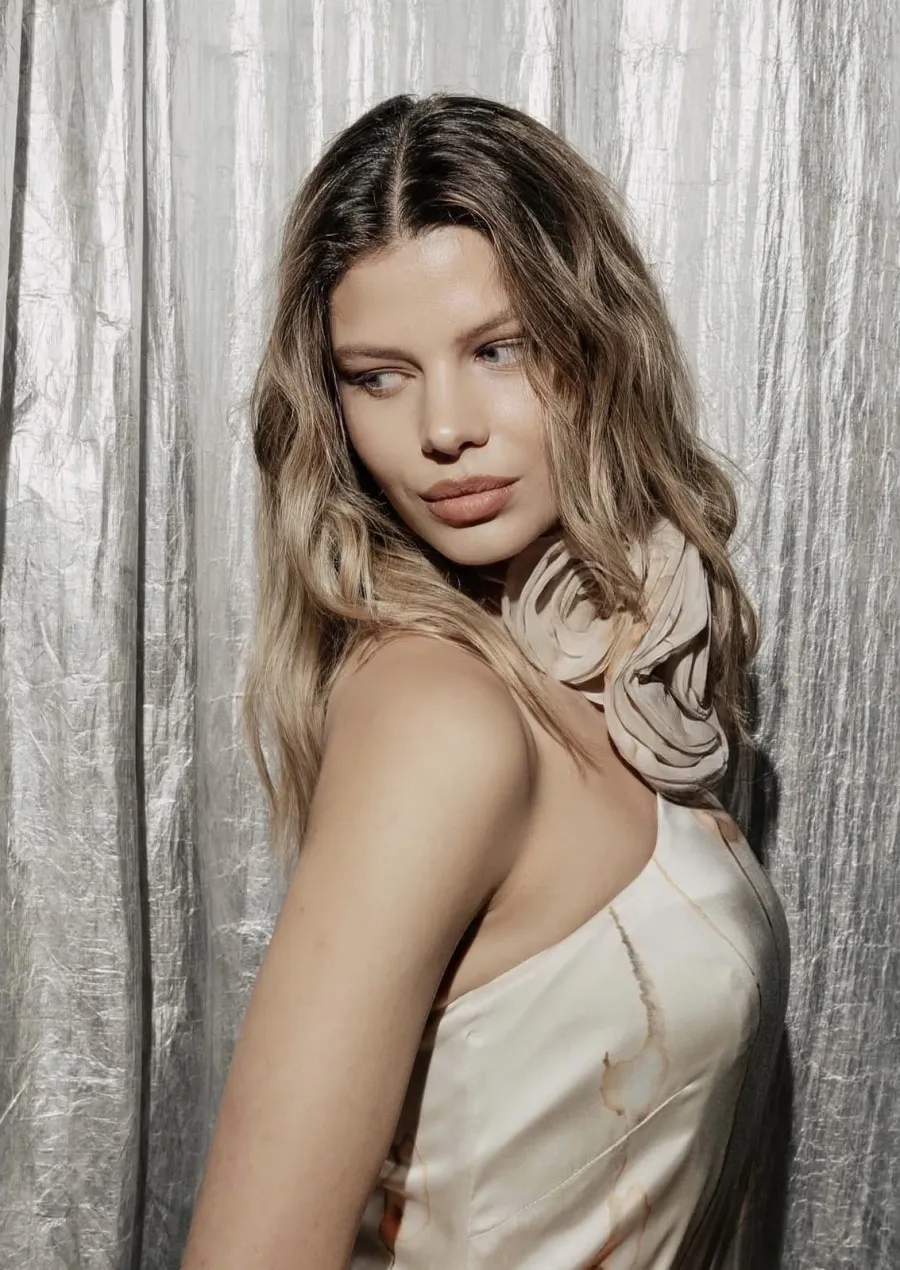Model with long blonde waves looking over her shoulder, in a cream satin top with a silk rose, in front of crinkled silver foil.