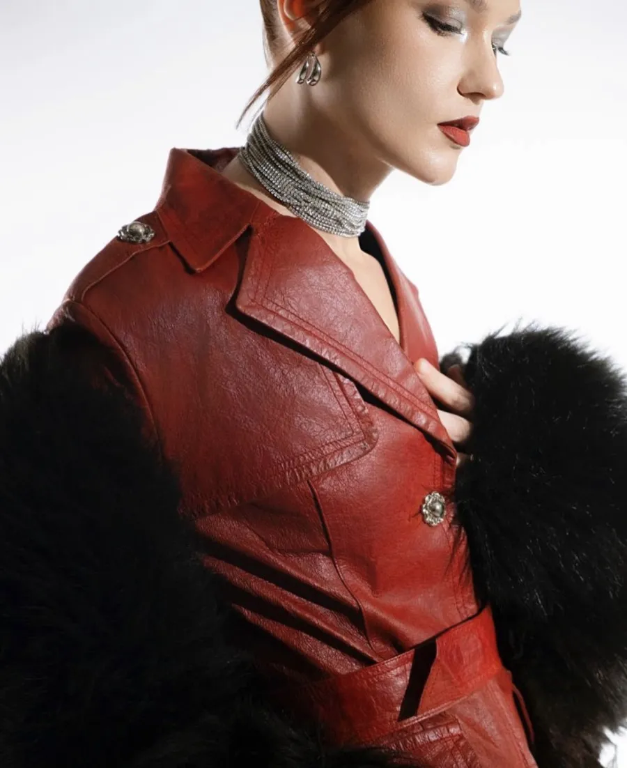 Model with auburn hair pinned up, in a red leather trench coat, black fur and a silver choker, eyes lowered, against a white studio backdrop.