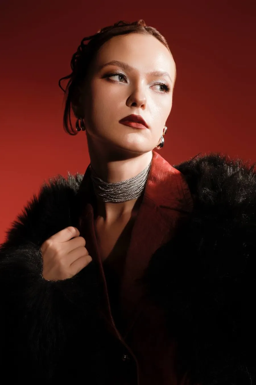 Model with auburn hair in a black fur coat and silver choker with dark red lips, against a deep red backdrop.