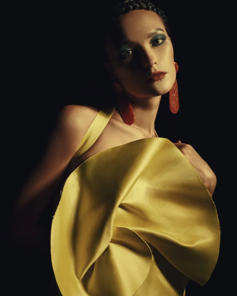 Model in a sculptural yellow satin dress and long red earrings, lit from the side against black.