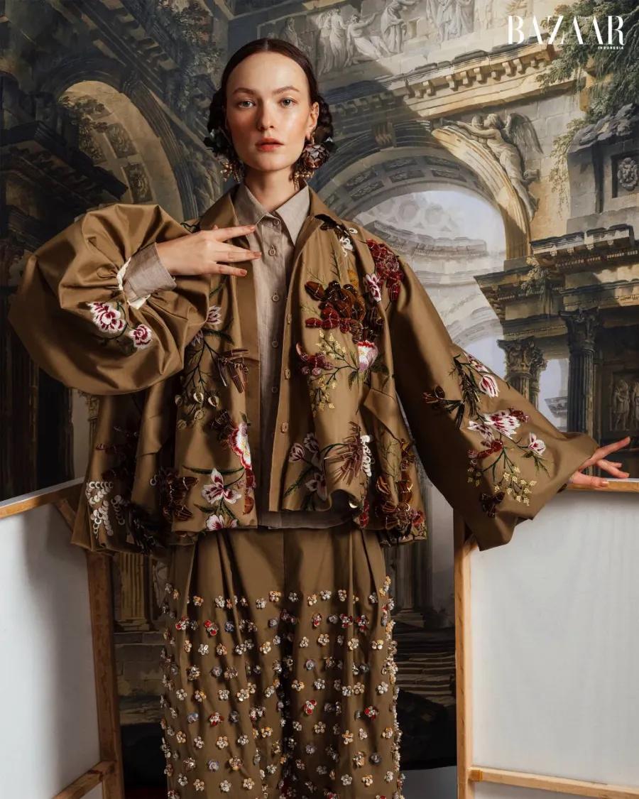 Model in an embroidered olive jacket with floral appliqué and beaded trousers, arms spread, in front of a painted classical backdrop.