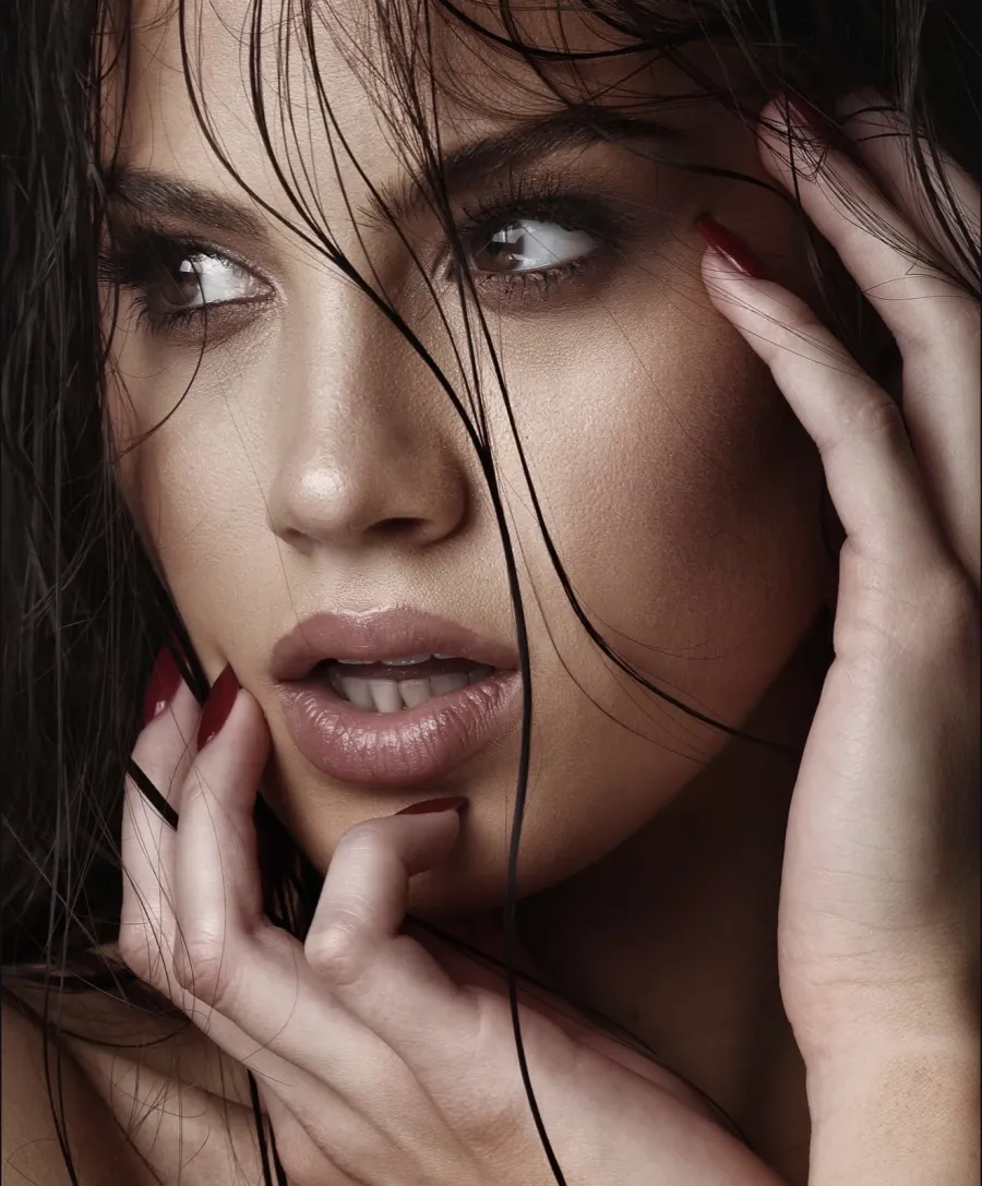 Beauty close-up of a dark-haired model with wet hair across her face, hands at her cheeks, glancing sideways.